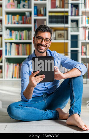 Portrait of barefoot young man sitting in front of bookshelves on the floor using digital tablet Stock Photo
