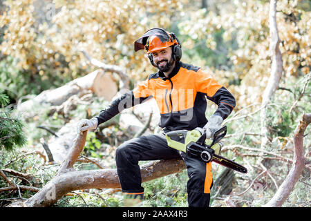 Portrait of a professional lumberman in protective workwear sitting with a chainsaw on the felled tree, resting after the hard work in the forest Stock Photo