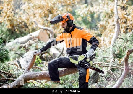 Portrait of a professional lumberman in protective workwear sitting with a chainsaw on the felled tree, resting after the hard work in the forest Stock Photo
