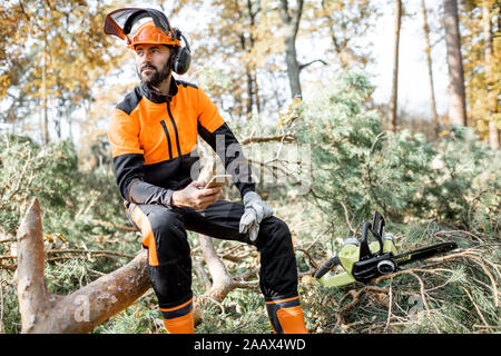 Portrait of a professional lumberman in protective workwear sitting with mobile phone on the felled tree, resting after the hard work in the forest Stock Photo