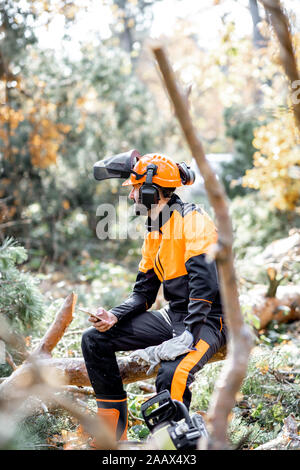 Portrait of a professional lumberman in protective workwear sitting with a chainsaw on the felled tree, resting after the hard work in the forest Stock Photo
