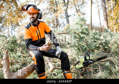 Portrait of a professional lumberman in protective workwear sitting with mobile phone on the felled tree, resting after the hard work in the forest Stock Photo