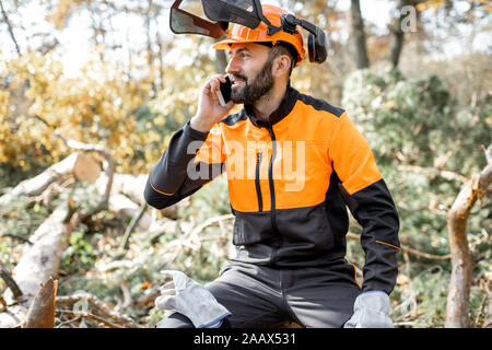 Professional lumberman in protective workwear talking on phone, while sitting on the felled tree, resting after the hard work in the forest Stock Photo