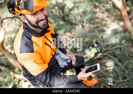 Portrait of a professional lumberman in protective workwear sitting with mobile phone on the felled tree, resting after the hard work in the forest Stock Photo
