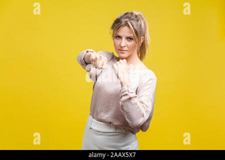 Portrait of brave confident young woman with blonde hair in casual beige blouse standing, holding clenched fists up ready to boxing, fighting spirit. Stock Photo