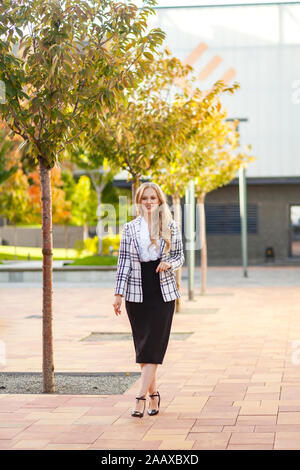 Full length portrait of stunning beautiful positive woman, confident businesslady with long blond hair wearing plaid jacket and skirt walking alone on Stock Photo