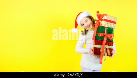 Cute little girl in a santa claus checker holds gift boxes in multi-colored wrapping papers and smiles cheerfully. Yellow studio background. Gifts con Stock Photo
