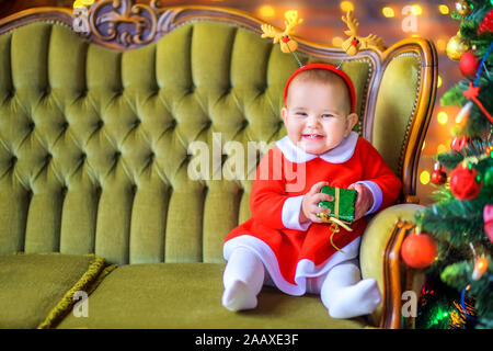 Cute little girl in a red dress is sitting on the sofa with a gift box in her hands and smiling. Christmas and New Year Stock Photo