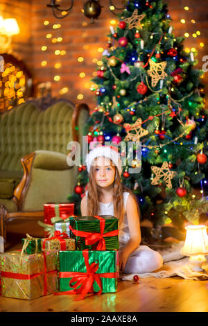 A cute girl in a white dress and Santa Claus hat is sitting on the floor with a large pile of gifts. Against the background of a festive tree. Christm Stock Photo