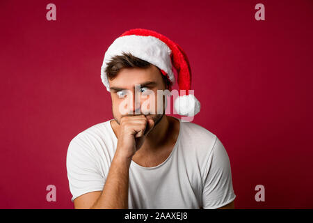 Young handsome caucasian guy in a white t-shirt and Santa hats stands on red background in studio and Sick after holiday food. Close up portrait europ Stock Photo