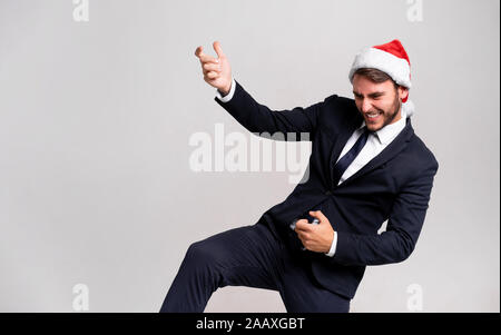 Young handsome caucasian guy in business suit and Santa hats stands on white background in studio Plays on an imaginary guitar. Close up portrait busi Stock Photo