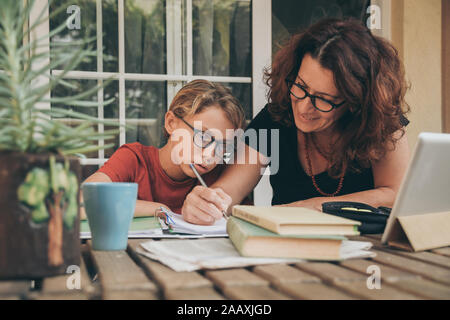 Tired student doing homework at home with school books, newspaper and digital pad helped by his mother. Mum writing on the copybook teaching his son. Stock Photo