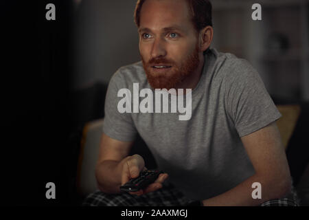 Portrait of bearded adult man watching TV at night while siting on couh in dark room and switching channels, copy space Stock Photo