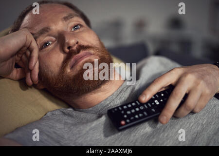 Portrait of bearded adult man watching TV at night while lying on couh in dark room and switching channels, copy space Stock Photo