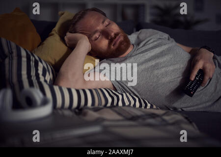 Portrait of bearded adult man sleeping on sofa while watching TV at night, copy space Stock Photo
