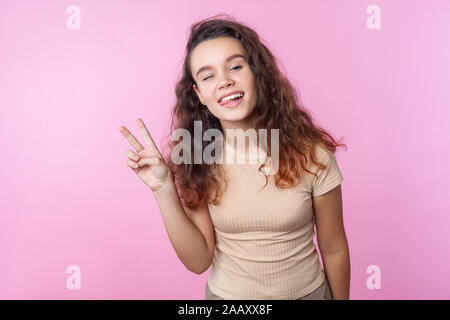 Portrait of cheerful vivid teenage girl with long curly brunette hair wearing casual style beige clothes winking at camera and showing victory gesture Stock Photo