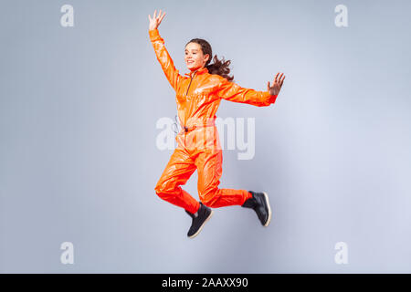 Full length image of excited energetic teen girl with brunette hair wearing bright orange jumpsuit dancing flying or jumping in the air with excited f Stock Photo