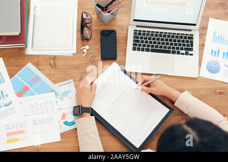 Above view background of unrecognizable businesswoman working at wooden desk with papers and documents laid out and word Communication on laptop screen, copy space Stock Photo