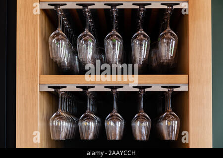Closeup many upside down empty clear transparent crystal upturned wine glasses hanging in straight rows on brown wooden shelf, rack, showcase, stainle Stock Photo