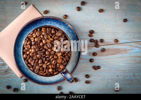 Flat lay cappuccino cup with coffee beans scattered on table, blue coffee cup top view closeup on wooden background in sun light. Cafe and bar, barist Stock Photo
