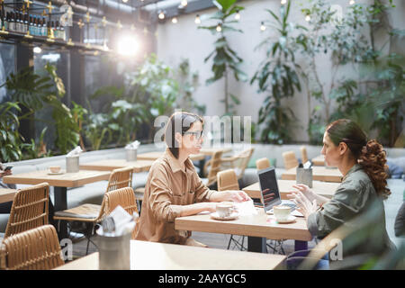 Side view portrait of two modern young women sitting at table in cafe discussing business during meeting on outdoor patio, copy space Stock Photo