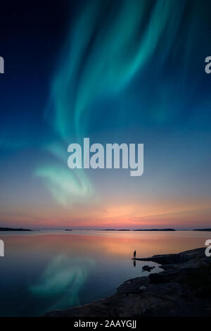 Silhouette of a man standing by a lake shore and looking at a beautiful aurora borealis on the sky with reflections on the calm lake. Stock Photo