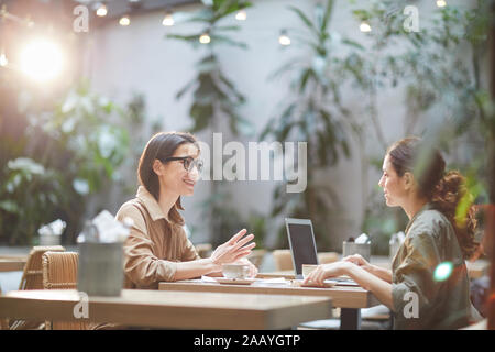 Side view portrait of two modern young women sitting at table in cafe and smiling cheerfully enjoying lunch together, copy space