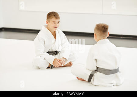Two boys in kimono sitting on the floor opposite each other and talking before training in gym Stock Photo