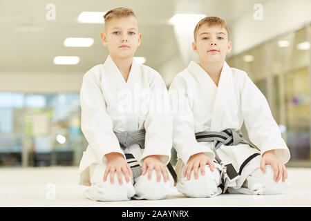 Portrait of two little boys in white kimono sitting on the floor and looking at camera they doing karate in gym Stock Photo