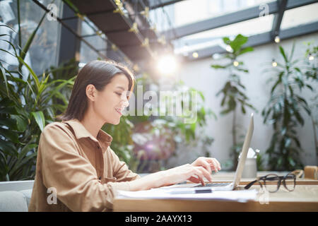 Side view portrait of smiling young woman using laptop in outdoor cafe terrace decorated with plants, copy space Stock Photo