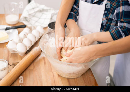 Hands of young woman and her little son kneading selfmade dough in bowl while both standing by table in the kitchen Stock Photo