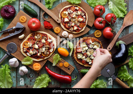 Vegetarian homemade festive food. Woman hand pouring grated cheese on pizzas with mushrooms, bell peppers, eggplants and goat cheese and raw ingredien Stock Photo
