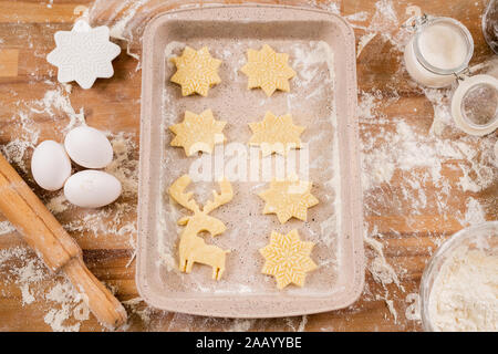Flatlay of tray with two rows of raw cookies surrounded by eggs, rolling pin, flour, cutters and open jar of sugar Stock Photo