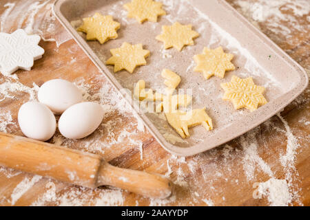 Tray with two rows of raw Christmas cookies with three eggs, rolling pin, flour and cutters near by on table Stock Photo