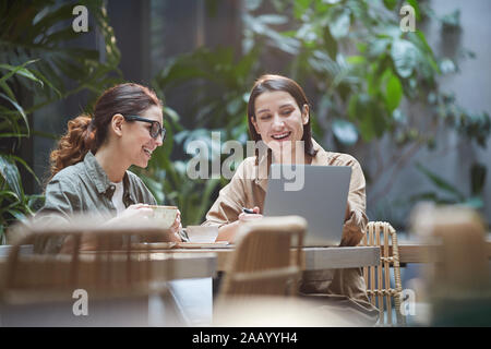 Portrait of two young women laughing happily while using laptop on outdoor cafe terrace decorated with plants, copy space Stock Photo