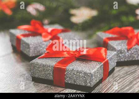 Three silver Christmas Gift boxes with red ribbon tied in a bow around the, sitting in background are the branches of a Christmas tree & decorations Stock Photo