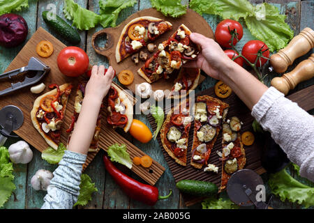 Vegetarian homemade party food concept. Woman and child hands reaching for pizza with champignons, bell peppers, eggplants and goat cheese with raw ve Stock Photo