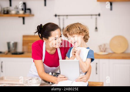 Joyful boy and his mother in aprons laughing at video in touchpad while sitting in the kitchen and choosing what to cook Stock Photo