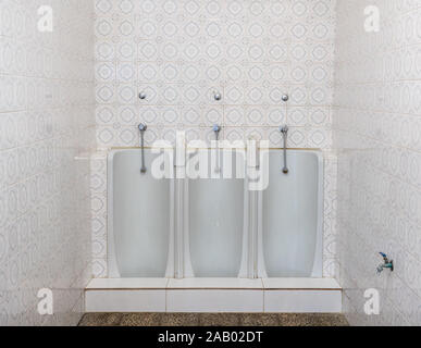 Set of three side by side full length mens urinals in porcelain against a tiled wall in small restroom Stock Photo
