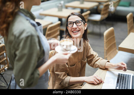 High angle portrait of young woman smiling at waitress while working at cafe table, copy space Stock Photo