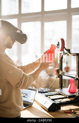 Young businessman in vr headset holding red plastic object in front of himself while going to put it on 3d printer in office Stock Photo