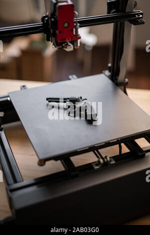 Black revolver gun lying on working surface of modern 3d printer with printhead hanging above Stock Photo