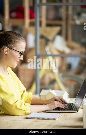 Happy young architect or engineer in casual yellow pullover sitting by table in front of laptop and browsing in the net for data Stock Photo