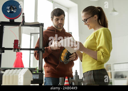 Two young contemporary engineers consulting about new creative project while one of them making notes in notepad Stock Photo