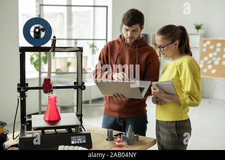 Two young creative designers searching for new online ideas for creative project while standing in front of 3d printer Stock Photo