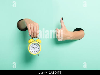 Female hand hold alarm clock and show tumb sign through a hole on neon mint background. Just in time concept. Stock Photo