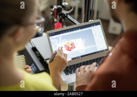 One of two young architects or designers pointing at cube sketch on laptop display during discussion of its details Stock Photo