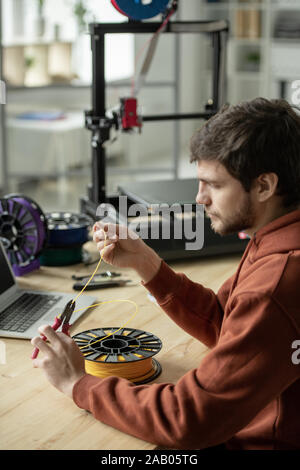 Creative man sitting by table and cutting piece of yellow filament while going to make 3d printing of new object Stock Photo