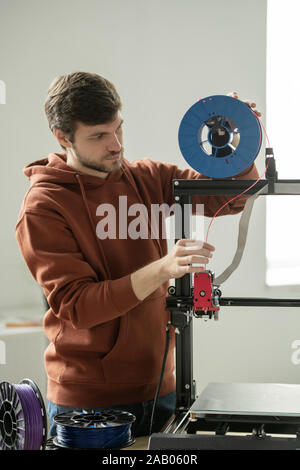 Young man in casualwear putting red filament into printhead of 3d printer before printing new items Stock Photo