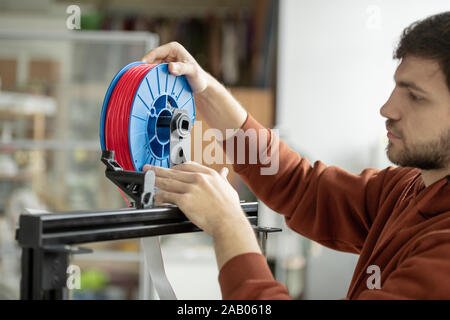 Young designer putting spool with red filament in 3d printer while going to print new items at work Stock Photo